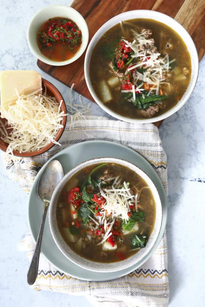 Two bowls of Italian lentil soup with beef, vegetables, and greens, topped with shredded cheese and red chili garnish. A bowl of shredded cheese, a small dish of chili sauce, and a spoon are also visible on a striped cloth and wooden surface.