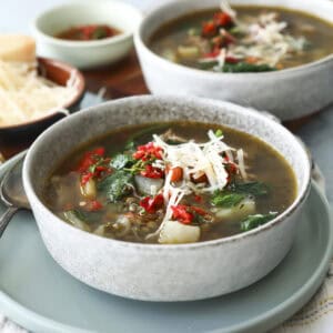 A bowl of Italian lentil soup topped with shredded cheese and chopped red peppers sits on a plate, with another bowl of soup and a small dish of grated cheese in the background.