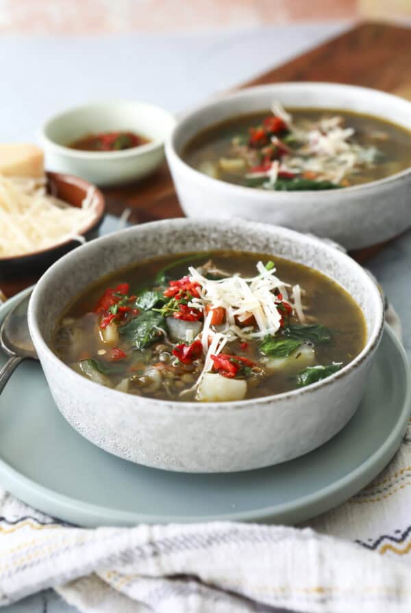 A bowl of Italian lentil soup topped with shredded cheese and chopped red peppers sits on a plate, with another bowl of soup and a small dish of grated cheese in the background.