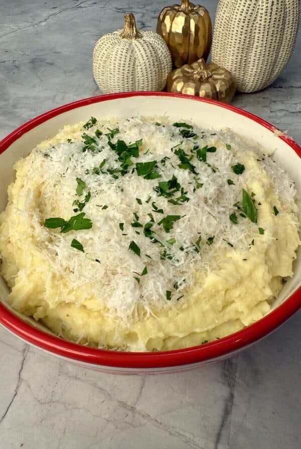 A bowl of roasted garlic parmesan mashed potatoes topped with shredded cheese and chopped herbs sits on a marble surface, with decorative white and gold pumpkins in the background.