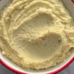 A bowl of creamy mashed potatoes with a smooth, swirled surface, served in a white bowl with a red rim on a light marble background.