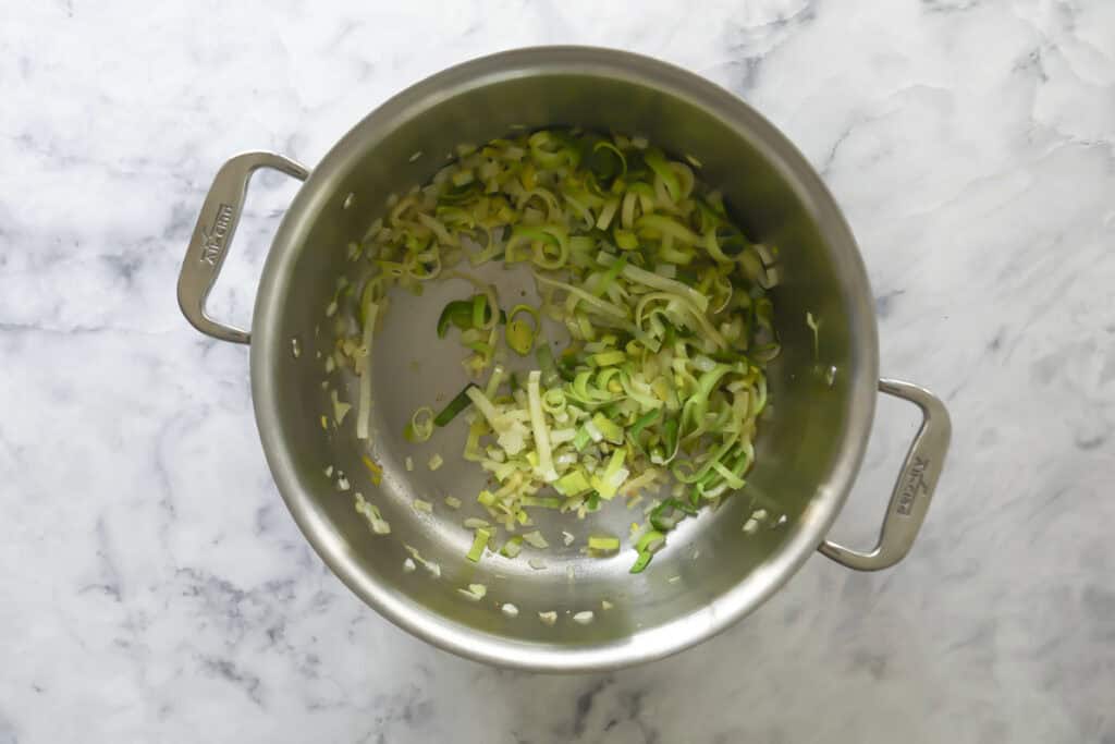 A stainless steel pot on a marble countertop containing sliced leeks that are being sautéed. The leeks are lightly cooked and spread across the bottom of the pot.
