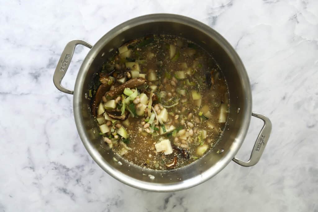 A stainless steel pot filled with vegetable soup containing beans, leafy greens, and chopped vegetables sits on a white marble surface.