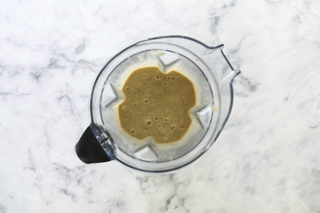 A top-down view of a blender containing a creamy light brown mixture, placed on a white marble surface.