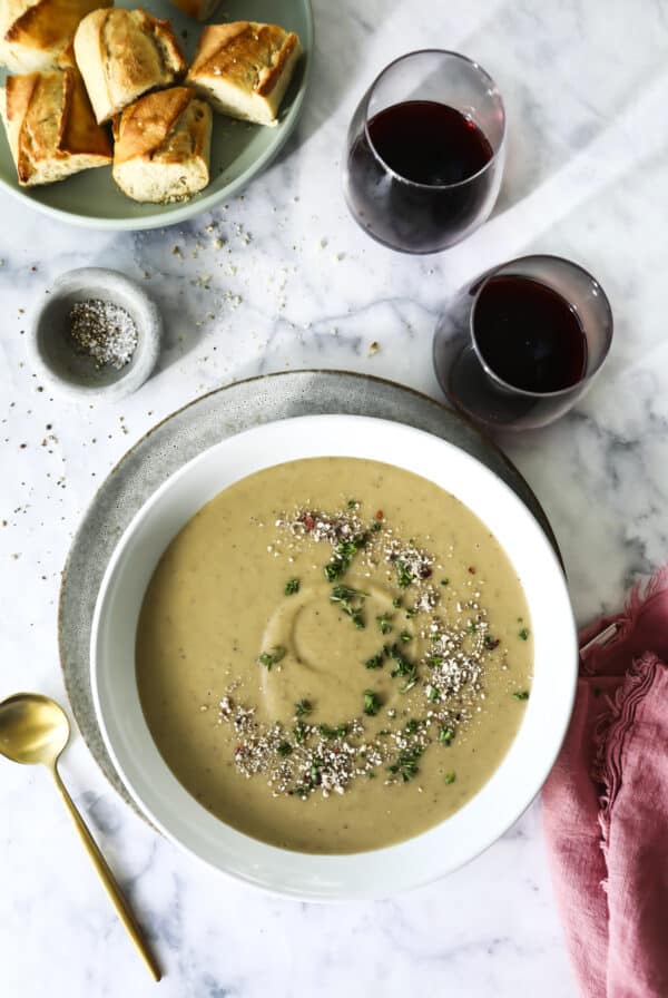A bowl of vegan cream of mushroom soup topped with chopped herbs and pepper sits on a plate, next to a gold spoon, a pink napkin, two glasses of red wine, bread slices on a plate, and a small bowl of pepper, all on a marble surface.