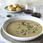 A bowl of vegan cream of mushroom soup garnished with herbs and seeds sits on a plate, with two glasses of red wine and a plate of bread rolls in the background on a marble surface.