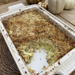 A baked Potato Gattò casserole with a golden, crispy breadcrumb topping in a white rectangular dish, partially served. Decorative pumpkins are on the wooden table in the background.