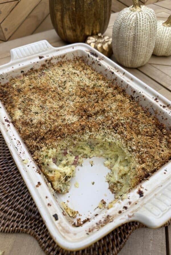 A baked Potato Gattò casserole with a golden, crispy breadcrumb topping in a white rectangular dish, partially served. Decorative pumpkins are on the wooden table in the background.