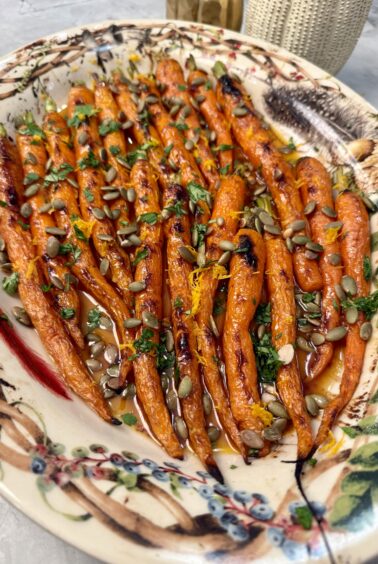 A decorative platter of roasted carrots garnished with chopped herbs, orange zest, and sunflower seeds, arranged neatly in rows on a colorful, patterned plate.