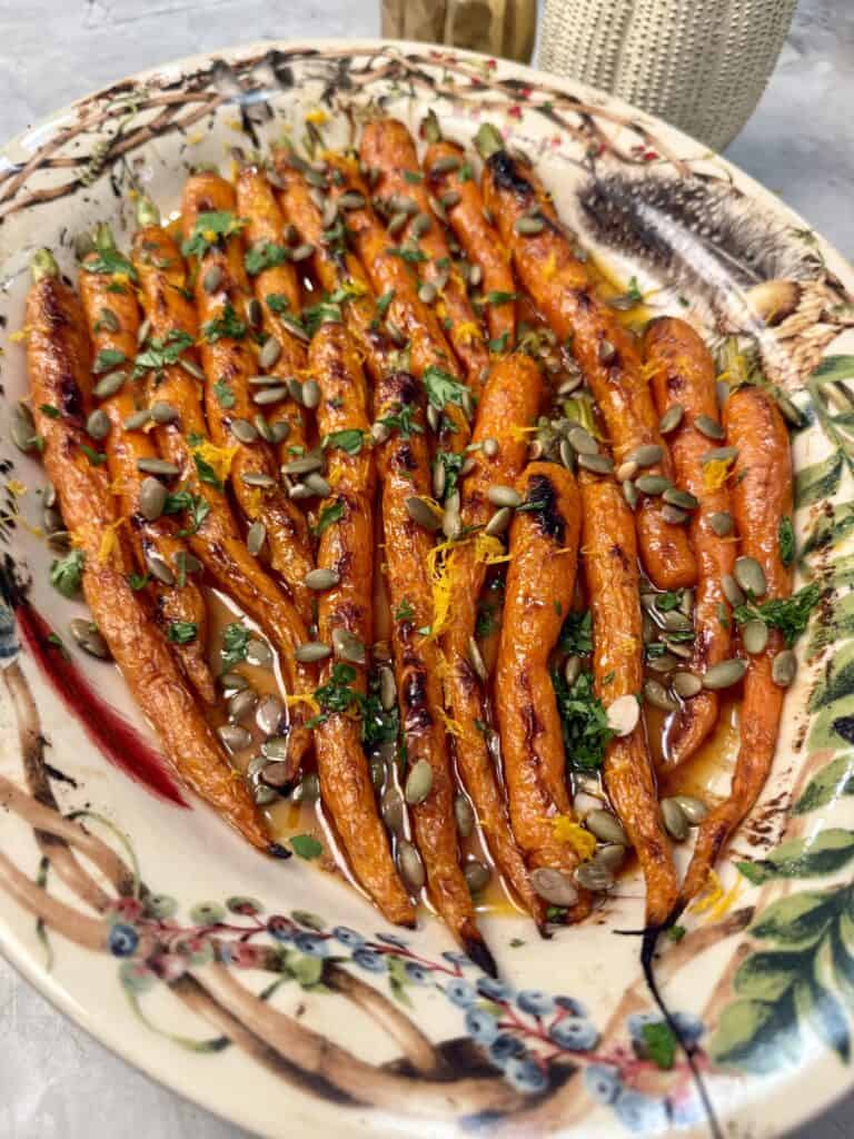 A decorative platter of roasted carrots garnished with chopped herbs, orange zest, and sunflower seeds, arranged neatly in rows on a colorful, patterned plate.