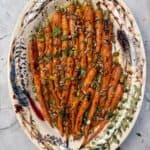 A decorative oval plate with chipotle honey roasted carrots topped with chopped herbs, pumpkin seeds, and a drizzle of glaze, set on a marble surface.