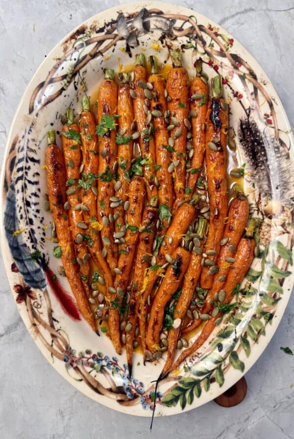 A decorative oval plate with chipotle honey roasted carrots topped with chopped herbs, pumpkin seeds, and a drizzle of glaze, set on a marble surface.