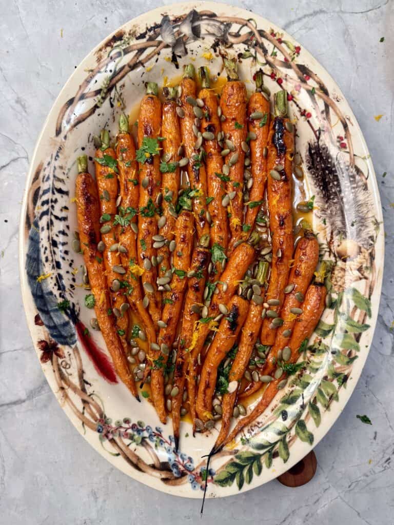 A decorative oval plate with chipotle honey roasted carrots topped with chopped herbs, pumpkin seeds, and a drizzle of glaze, set on a marble surface.