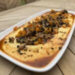 A rectangular white dish filled with sausage and polenta. The creamy yellow polenta, topped with a savory mixture of ground meat, diced orange vegetables, herbs, and a rich red sauce, set on an outdoor wooden table.
