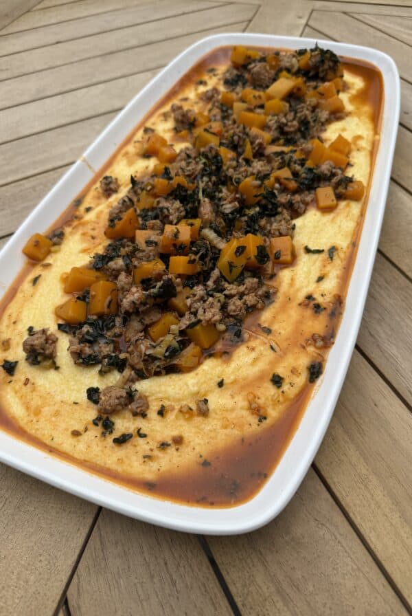 A rectangular white dish filled with sausage and polenta. The creamy yellow polenta is topped with a savory mixture of ground sausage, diced orange and green vegetables, herbs, and a reddish sauce, placed on a wooden table.
