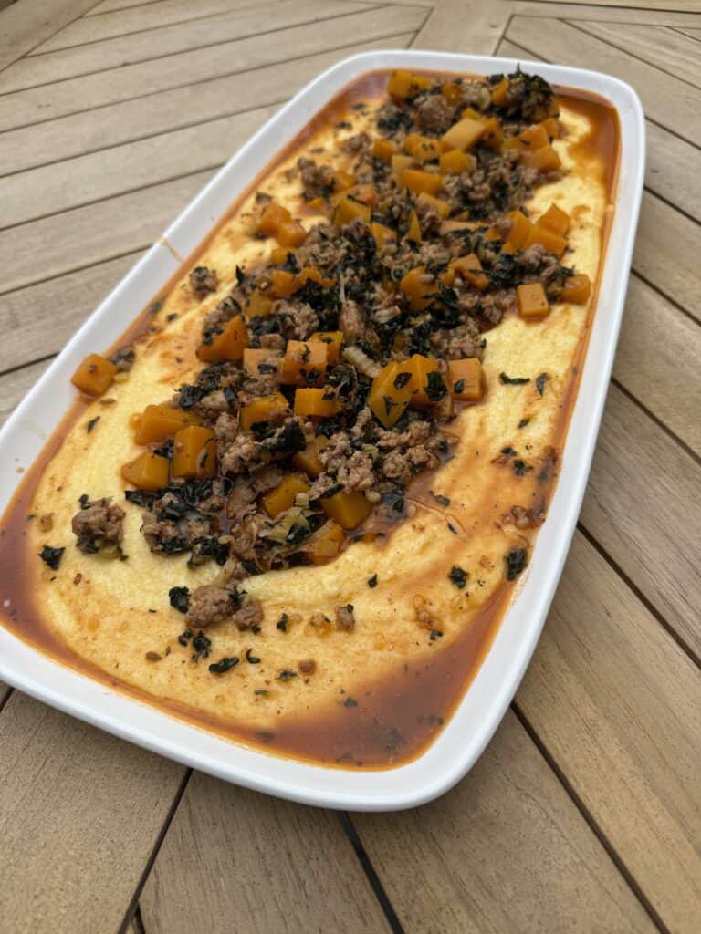 A rectangular white dish filled with sausage and polenta. The creamy yellow polenta is topped with a savory mixture of ground sausage, diced orange and green vegetables, herbs, and a reddish sauce, placed on a wooden table.