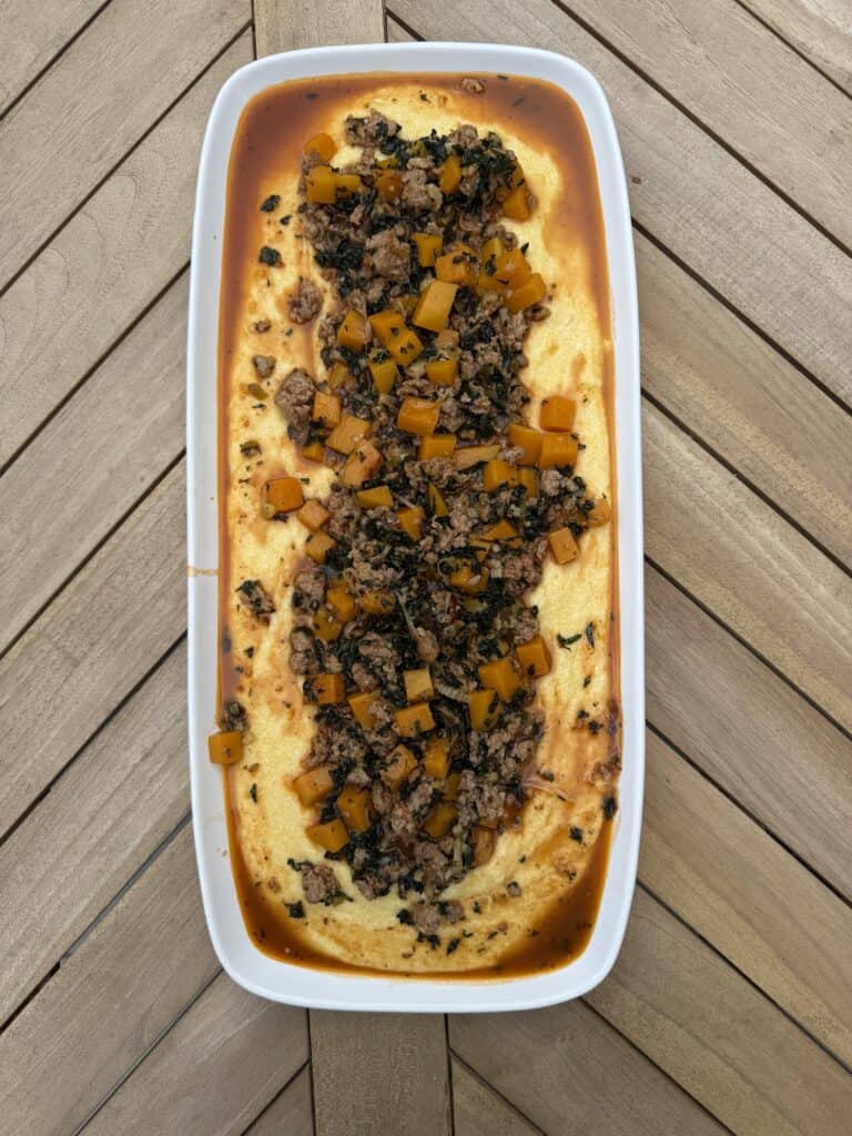 A rectangular white dish filled with sausage and polenta. The creamy polenta, topped with a savory mixture of ground meat, diced yellow squash, chopped greens, and a reddish sauce, placed on a herringbone-patterned wooden table.