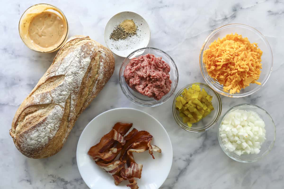 A loaf of bread, cooked bacon strips, ground beef, shredded cheddar cheese, diced pickles, diced onions, seasonings, and a bowl of sauce are arranged on a marble surface.