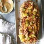 A toasted loaf of bread topped with melted cheese, chopped bacon, sauce, and sliced pickles on a rectangular plate, with a bowl of sauce and a spoon beside it on a textured surface.
