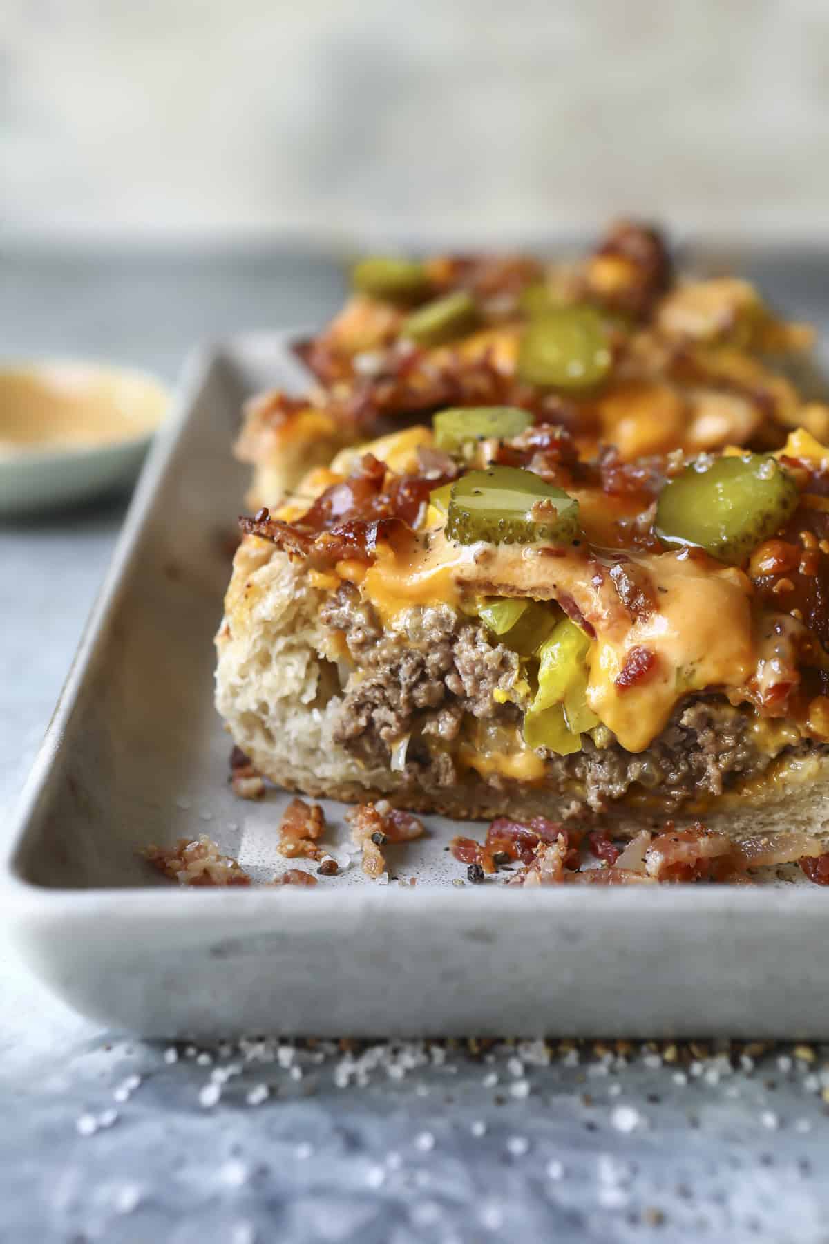 A close-up of a cheeseburger casserole topped with melted cheese, pickles, and crispy bacon bits, served on a rectangular baking dish. A small dish of sauce is blurred in the background.