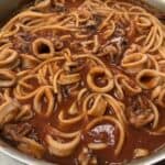 Spaghetti with calamari rings in a rich red tomato sauce, served in a stainless steel pan on a marble countertop.
