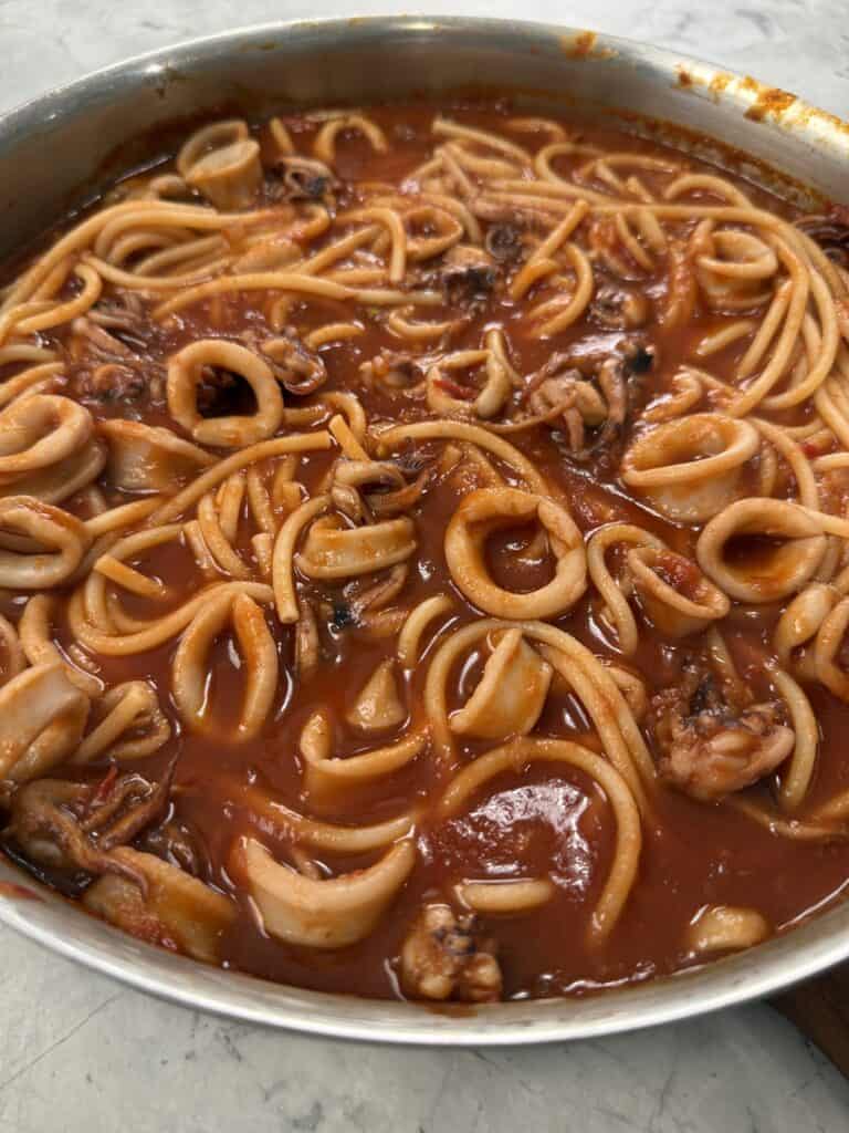 Spaghetti with calamari rings in a rich red tomato sauce, served in a stainless steel pan on a marble countertop.