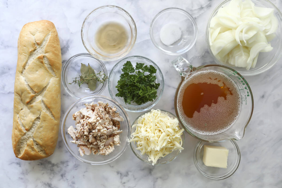 A top-down view of ingredients on a marble surface, including a loaf of bread, chopped chicken, shredded cheese, sliced onions, broth, butter, parsley, bay leaves, thyme, vinegar, sugar, and several empty glass bowls.