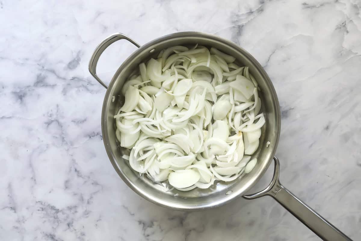 A stainless steel pan filled with sliced raw white onions sits on a white marble countertop.