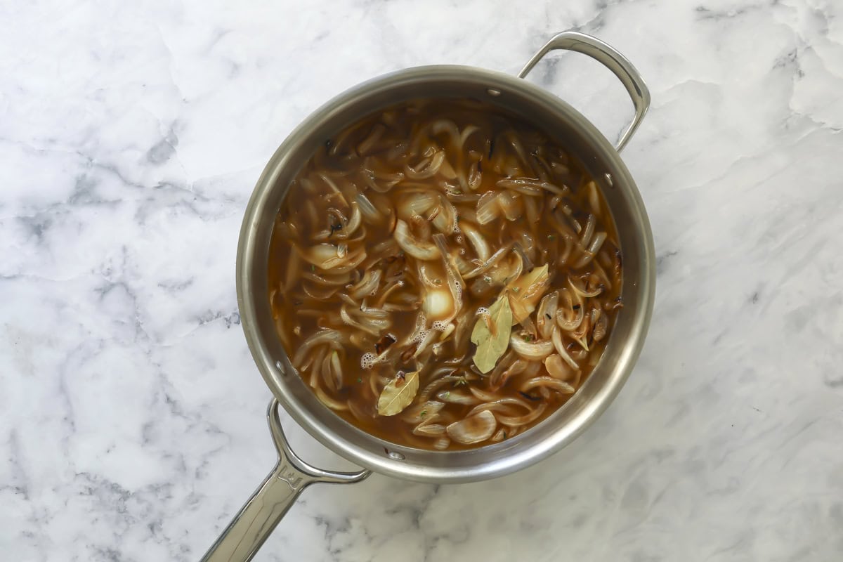 A stainless steel pot filled with sliced onions simmering in brown broth, topped with bay leaves, sits on a white marble surface.