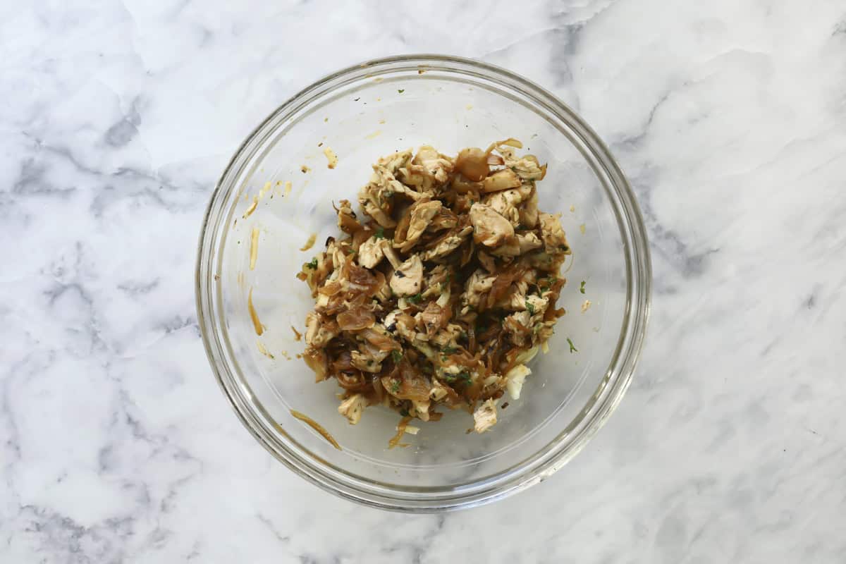 A glass bowl filled with shredded cooked chicken mixed with herbs and a dark sauce sits on a white marble surface.