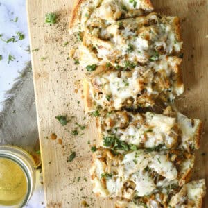 A wooden cutting board with slices of cheesy, herb-topped bread garnished with chopped parsley. Glasses of light-colored drink and a cup of soup are nearby, with a cloth napkin in the background. French Onion and Chicken Stuffed Bread