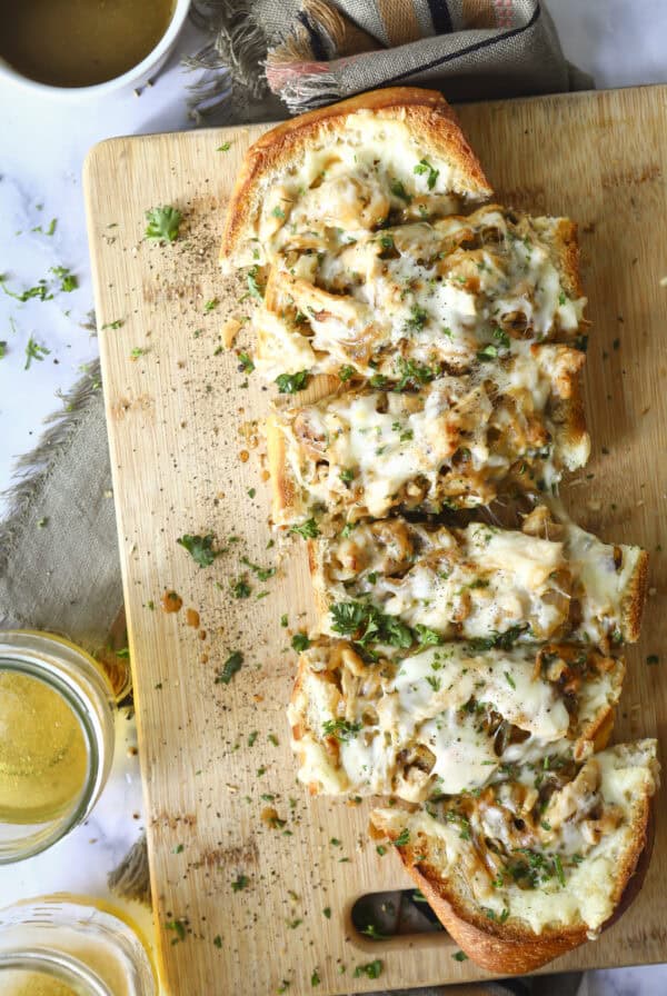 A wooden cutting board with slices of cheesy, herb-topped bread garnished with chopped parsley. Glasses of light-colored drink and a cup of soup are nearby, with a cloth napkin in the background. French Onion and Chicken Stuffed Bread
