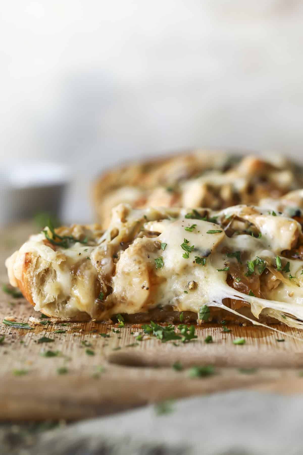 Close-up of a cheesy, golden-brown slice of bread topped with melted cheese, herbs, and pieces of cooked chicken, served on a wooden board, with fresh chopped parsley sprinkled on top.