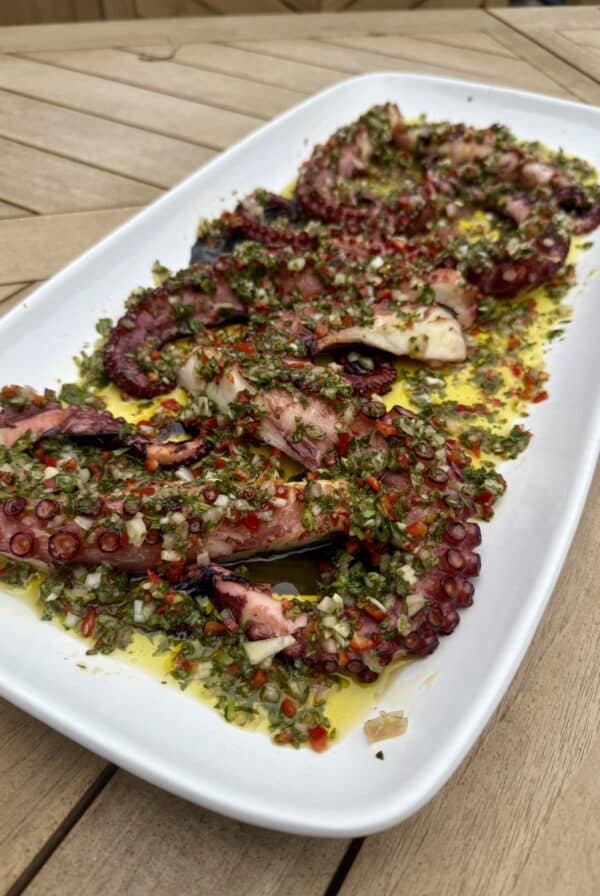 Grilled octopus tentacles topped with a fresh green herb sauce, served on a white rectangular plate, placed on a wooden table. The dish is garnished with chopped herbs, garlic, and red chili.