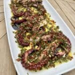 Grilled octopus tentacles served on a white rectangular plate, garnished with a vibrant mix of chopped herbs, red chili, and olive oil, placed on a wooden table.
