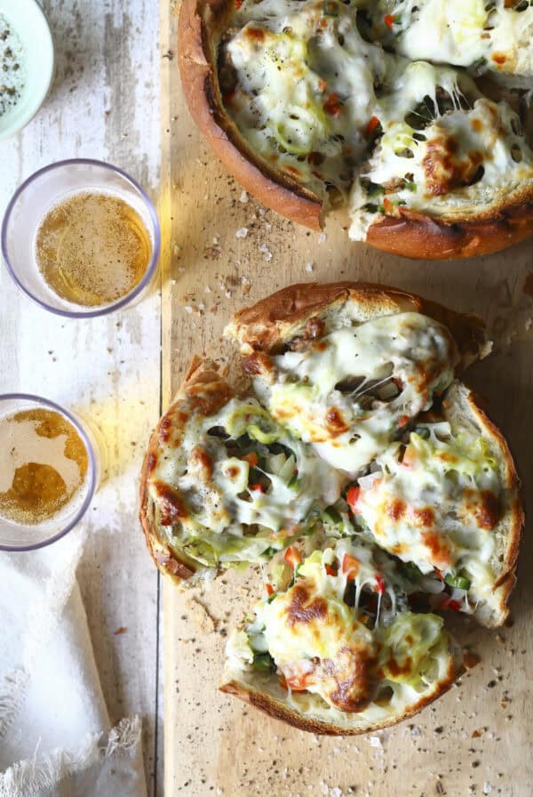 Two round bread bowls filled with melted cheese, vegetables, and herbs, sliced into portions on a wooden board make this Sausage and Pepper Stuffed Bread. Two glasses of beer sit nearby on a rustic white surface.