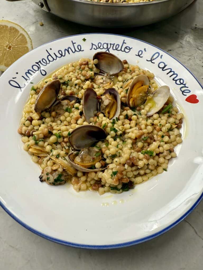 A plate of fregola pasta alle vongole (clams), garnished with herbs, sits on a white dish that reads “L’ingrediente segreto è l’amore” with a red heart, next to a halved lemon.