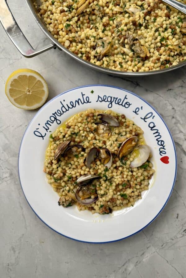 A plate of one pan pasta alle vongole (clams), garnished with herbs, sits on a marble surface. A lemon half is nearby, and the plate is inscribed with Italian text: L’ingrediente segreto è l’amore and a red heart.