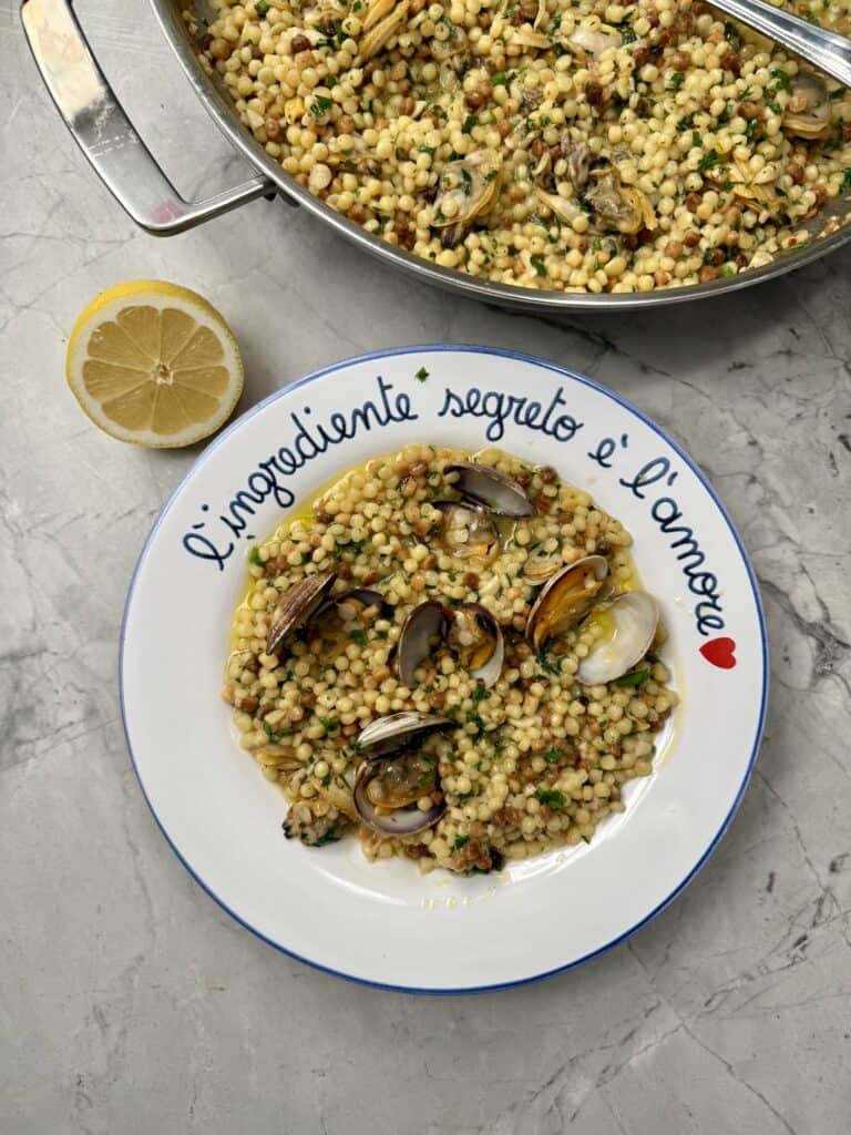 A plate of one pan pasta alle vongole (clams), garnished with herbs, sits on a marble surface. A lemon half is nearby, and the plate is inscribed with Italian text: L’ingrediente segreto è l’amore and a red heart.