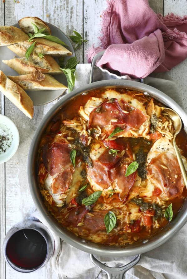 A skillet filled with Stuffed Chicken Dinner topped with prosciutto and basil in tomato sauce and orzo sits on a wooden table, next to sliced baguette, a glass of red wine, and a pink napkin.