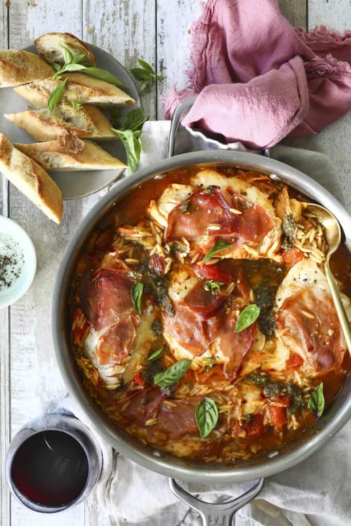 A skillet filled with Stuffed Chicken Dinner topped with prosciutto and basil in tomato sauce and orzo sits on a wooden table, next to sliced baguette, a glass of red wine, and a pink napkin.