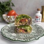 A Big Mac Lettuce wrap cut in half is stacked on a decorative green and white plate, showing layers of lettuce, tomato, ground beef, cheese, and sauce. Fresh vegetables and kitchen decor are in the blurred background.