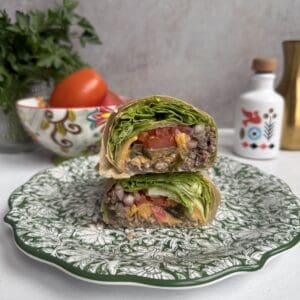 A Big Mac Lettuce wrap cut in half is stacked on a decorative green and white plate, showing layers of lettuce, tomato, ground beef, cheese, and sauce. Fresh vegetables and kitchen decor are in the blurred background.