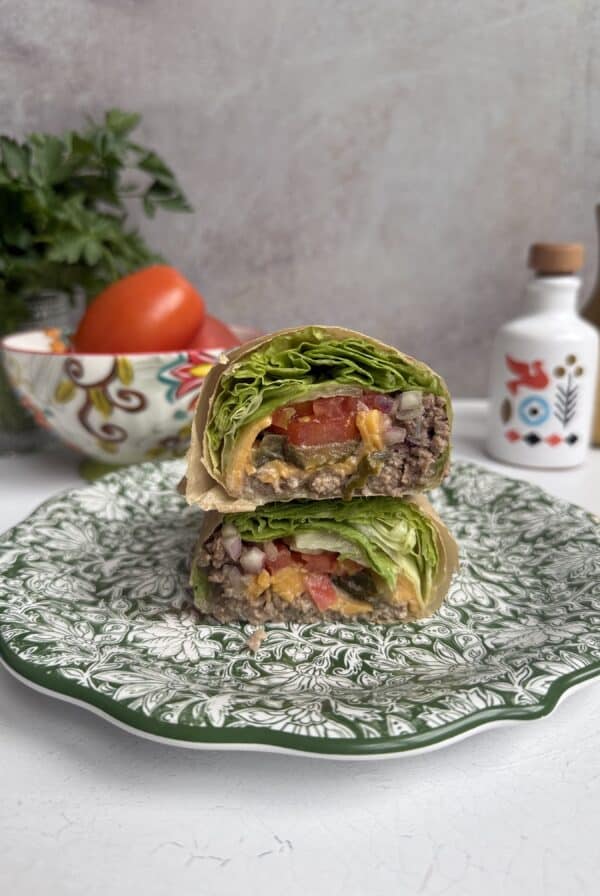 A Big Mac Lettuce wrap cut in half is stacked on a decorative green and white plate, showing layers of lettuce, tomato, ground beef, cheese, and sauce. Fresh vegetables and kitchen decor are in the blurred background.