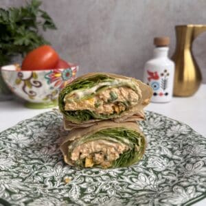 A Buffalo chicken lettuce wrap filled with greens and a creamy, orange-hued mixture is cut in half and stacked on a patterned green plate. In the background are a decorative bowl with tomatoes, a gold pitcher, and a white container.
