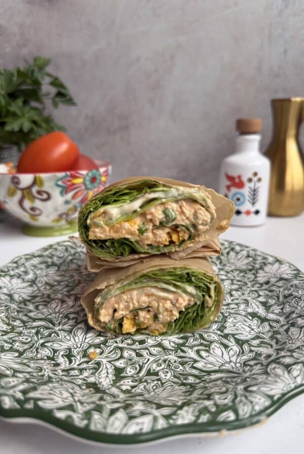 A Buffalo chicken lettuce wrap filled with greens and a creamy, orange-hued mixture is cut in half and stacked on a patterned green plate. In the background are a decorative bowl with tomatoes, a gold pitcher, and a white container.