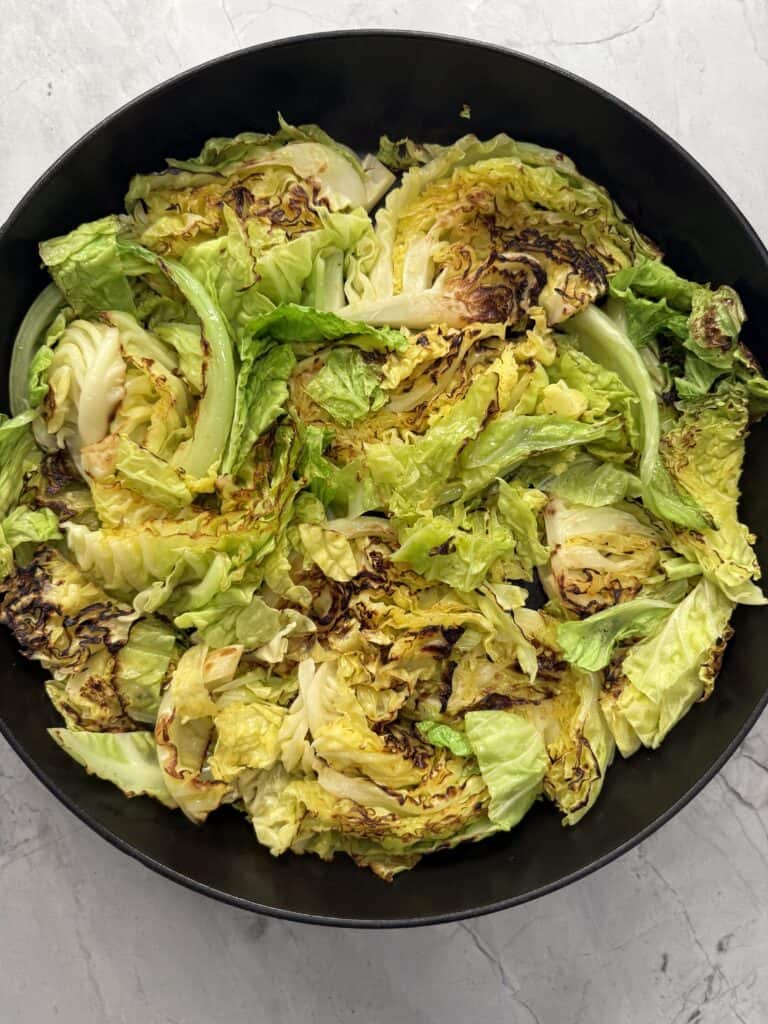 A black skillet filled with pan-seared green cabbage wedges, some leaves browned and caramelized like a deconstructed cabbage gratin, on a light gray marble surface.