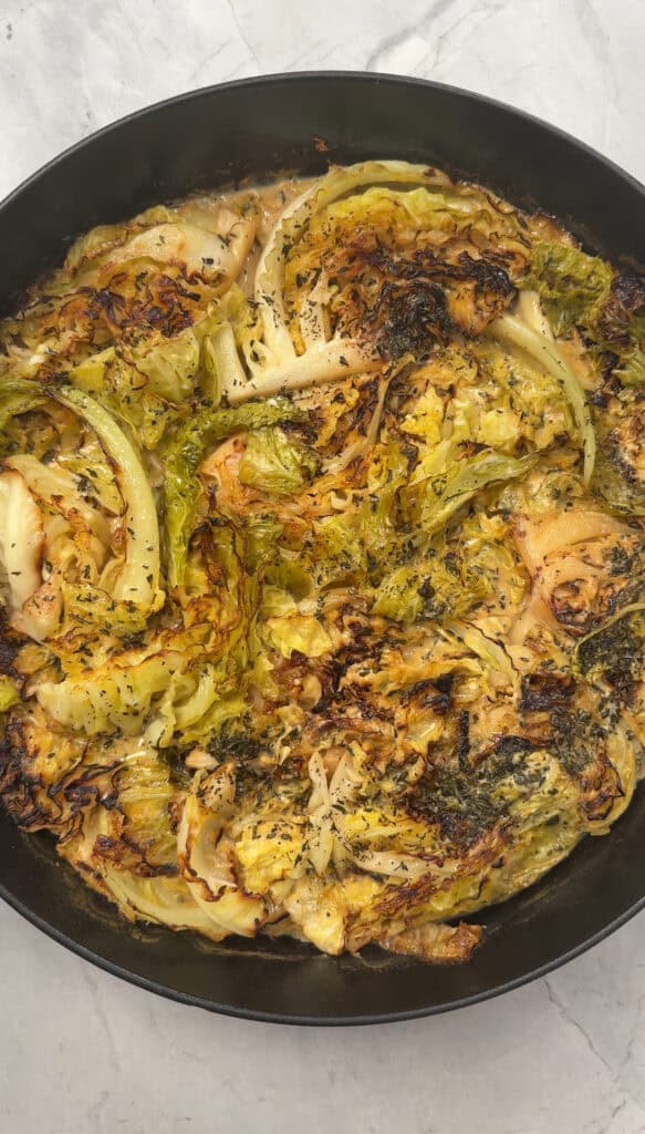 A round skillet filled with roasted cabbage gratin wedges, lightly charred on the edges and seasoned with herbs, sits on a light-colored surface.
