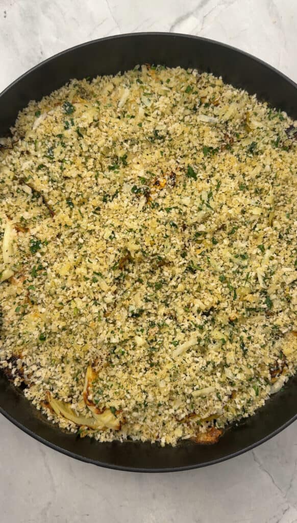 A round black baking dish filled with a golden-brown breadcrumb topping and chopped parsley, covering a baked cabbage gratin casserole on a light marble surface.