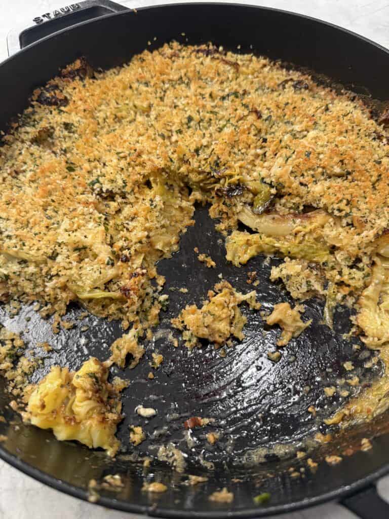 A black cast iron skillet with a golden-brown breadcrumb-topped cabbage gratin, partially served to reveal the crispy topping and tender cabbage gratin layers beneath.
